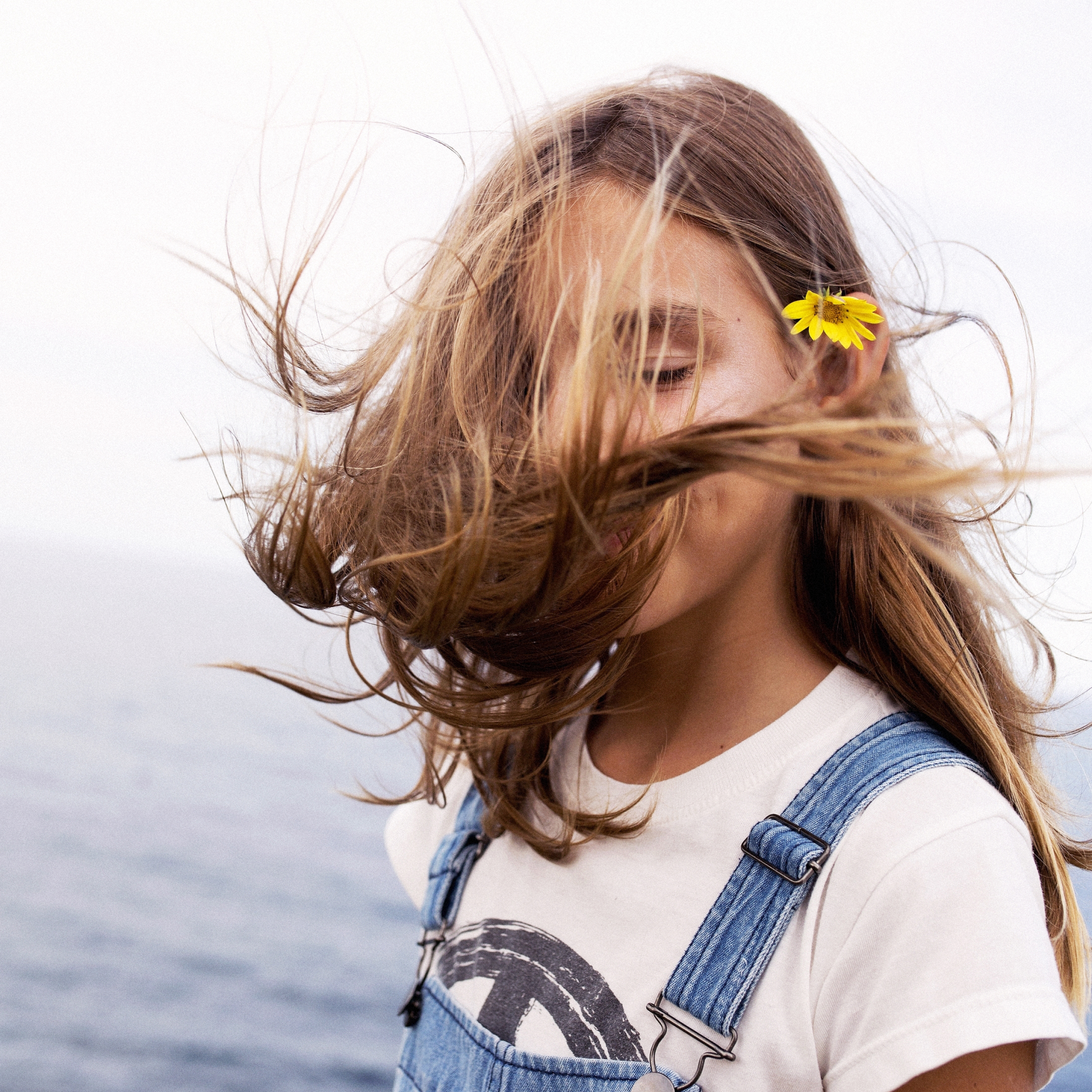 Zoomed in headshot of a young girl with eyes closed. Her brown hair is being blown by the wind and covers part of her face. A yellow flower is tucked behind her ear.